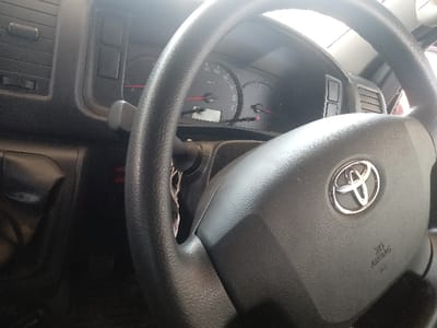The image depicts the interior of a vehicle, specifically the driver's perspective, located in Majengo ward, Kenya. The foreground is dominated by a black, textured steering wheel featuring a prominent silver Toyota emblem at its center. Below the emblem, the text "SRS AIRBAG" is visible. Behind the steering wheel, the instrument cluster is partially visible, displaying a speedometer calibrated up to at least 180 km/h, with needles at rest, indicating the vehicle is stationary or off. To the left of the instrument cluster, a vertical air vent is present on the dashboard, along with various dark gray plastic panels and controls. A control stalk extends from behind the steering wheel on the left side. No individuals, actions, or direct interactions are visible within the frame. The environment is the enclosed cabin of a vehicle, illuminated by natural light entering from an unseen source, suggesting daytime conditions.