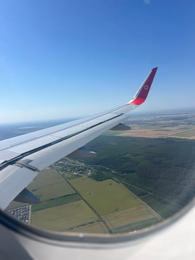An aerial view from an airplane window shows the wing of a commercial aircraft with a red and white livery featuring a pink heart logo. The wing extends across the frame, with the fuselage and a portion of the wing's upper surface visible. Below the wing, a landscape unfolds, characterized by a patchwork of green agricultural fields and dense forestation. A small town with scattered houses is visible nestled between the fields and the trees. In the distance, a hazy horizon suggests a body of water and a sprawling urban or semi-urban area. The sky is clear and bright blue. The scene depicts a flight over a rural and semi-urban area, potentially in the vicinity of Mogoșoaia, Romania, given the commonality of such landscapes and the presence of bodies of water often found near cities. There are no people or discernible actions captured in the image.