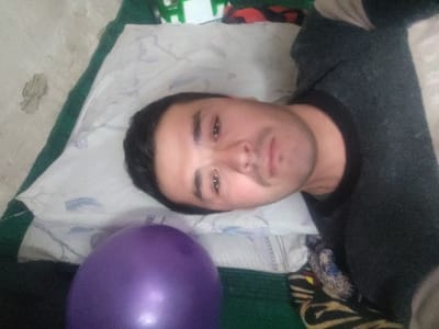 This image depicts an indoor scene in Yangikurgan, Uzbekistan, focused on one adult male lying down. The male, with short dark hair and light facial stubble, is positioned with his head resting on a white pillow with a subtle patterned design. He is wearing a dark grey, long-sleeved crew-neck garment. His gaze is directed towards the camera.
The subject is situated on a green mat or thin mattress. To the lower left of the frame, a large, inflated, spherical purple balloon is prominently visible, partially obscuring the mat. In the upper background, behind the subject's head, parts of other objects are discernible: a white and green item, a red and black patterned item, and a dark patterned fabric item. A light-colored, textured surface, possibly a wall or floor, is visible in the upper-left corner of the frame. The image is captured from a slightly elevated perspective, with the subject oriented sideways in the frame.