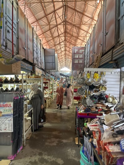 The image depicts the bustling interior of a market in Kara-Suu, Kyrgyzstan. The market is characterized by rows of stalls constructed from shipping containers, stacked and aligned to form a long, covered walkway. A corrugated metal roof, supported by a metal truss system, arches overhead, allowing natural light to filter into the space.

In the foreground, to the left, a display of kitchenware and utensils is visible, including metal strainers, ladles, and knives. Behind these, a stall features shelves laden with ceramic items, likely teapots and plates, and is being examined by a person wearing a grey headscarf and a long grey garment. Further down the aisle, another person, wearing a patterned orange dress and headscarf, walks alongside someone in a light pink outfit. They are moving away from the camera, deeper into the market.

The ground is a concrete surface, appearing somewhat damp and uneven. The overall impression is one of a vibrant, commercial environment filled with merchandise and shoppers. The stacked containers suggest a unique architectural approach to market design, potentially utilizing repurposed materials. The items on display indicate a mix of household goods and possibly local crafts.