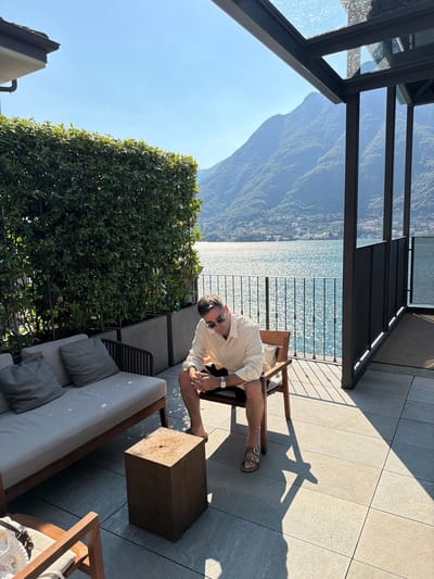 A man is seated on a wooden chair on an outdoor patio overlooking a lake and mountains. The man is wearing a light-colored button-down shirt, dark shorts, and sandals. He has sunglasses on and is looking down. To his left is a sofa with two cushions. In front of the sofa is a wooden cube-shaped table. The patio is paved with large gray tiles, and shadows from a pergola with a glass roof fall across the tiles. A manicured green hedge runs along the back of the patio. Beyond the hedge, a railing separates the patio from a shimmering blue lake. In the background, lush green mountains rise steeply from the far side of the lake, with a small town visible along the shoreline. The sky is clear and blue. The location is Pognana Lario, Italy.