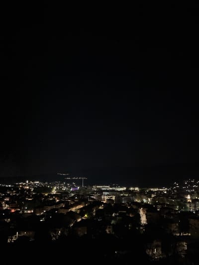 A night-time view in Sveti Konstantin, Bulgaria, shows a dark body of water bordered by the lit-up coastline. The coast is lined with buildings, illuminated trees, and streetlights. The lights create a dense, urban illumination pattern. No people are visible, only the urban environment.