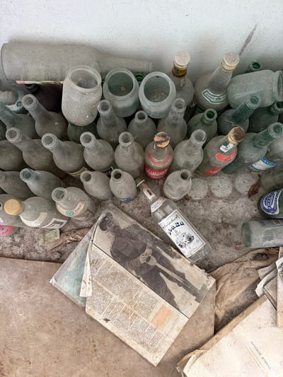 In Nikolsky, Russia, a collection of dusty glass bottles and jars are scattered across a concrete surface, possibly abandoned within a building or outdoor storage area. The bottles vary in size and color, with many appearing to be clear or green glass. Some bottles have caps or stoppers, while others are open. Several bottles have faded labels, suggesting they once contained liquids. Interspersed among the bottles are pieces of aged paper, including what appears to be a newspaper or magazine with a black and white photograph of a person in uniform, and a book with Cyrillic text. The overall environment is gritty and neglected, with dust and debris covering most surfaces.