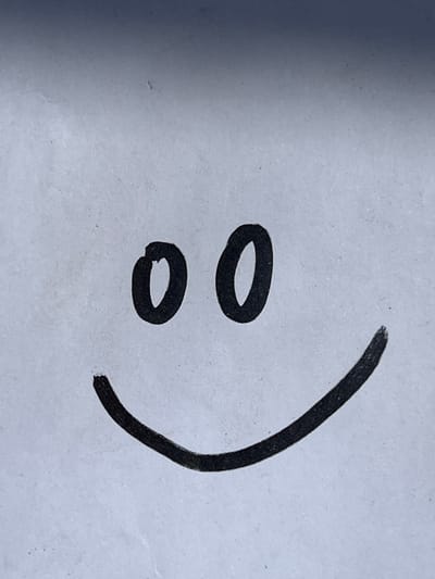 A simple smiley face is drawn on a white surface. The smiley face consists of two circular eyes and a curved line for a mouth. The drawing is in black ink. The background is a textured white surface with some subtle shading that creates a gradient from dark at the top to lighter at the bottom, possibly due to lighting. There is no discernible connection to the city of Lanžhot, Czechia.
