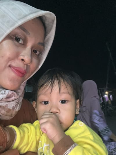 The image is a selfie taken at night, likely outdoors, in Wedarijaksa, Indonesia. In the foreground, a woman wearing a light-colored hijab and a pink and white patterned scarf is holding a young child. The woman has her face close to the camera, and her lips are painted red. The child, wearing a bright yellow shirt, has their fist near their mouth and is looking at the camera. In the background, another person wearing a dark purple hijab is partially visible, along with some streetlights and buildings. The overall lighting suggests an urban or semi-urban setting at night.