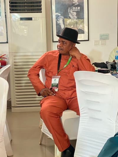 A medium, indoor shot depicts a man wearing a black hat and an orange jumpsuit. He is sitting in a white chair covered with a white cloth. He is wearing a lanyard with a name tag that reads "MID TERM REVIEW, PARTICIPANT." He is holding a pen in his right hand and resting his left hand on his neck. To his left is a white air conditioning unit and a framed photograph on the wall. A fire extinguisher is visible near the bottom left corner of the image. The location is presumed to be in Kwarau, Nigeria.