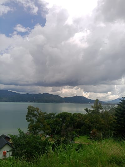 The image captures a panoramic view of Lake Toba in Kalalengkio, Indonesia, under a dramatic sky filled with heavy, grey clouds. The vast, calm body of water occupies the majority of the frame, reflecting the subdued light. Across the lake, a range of verdant, tree-covered mountains forms a distant backdrop. Closer to the foreground, lush green vegetation, including tall grass, dense shrubs, and scattered trees, dominates the landscape. A few buildings, including a white house with a dark roof and an orange water tank, are visible nestled amongst the foliage on the left side of the image. A white vehicle is parked on a grassy area further back, amidst the trees. The overall scene depicts a serene natural environment with hints of human habitation.