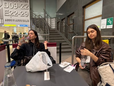This image depicts two young adult females seated at a dark table within a public area of the Milan Malpensa Airport, located in Somma Lombardo, Italy, identifiable by "IMBARCHI-GATES" and "ALDA MERINI" terminal signage.
The female on the left, with dark hair, wears a dark, buttoned top and is actively drinking from a clear plastic water bottle. A dark handbag is partially visible to her left. The female on the right, also with dark hair, wears a brown leather-like jacket over a light-colored top and dark pants; she holds a clear plastic water bottle in her left hand. A white plastic shopping bag with handles is centered on the table between them, along with crumpled white paper and a small wrapper near the right female. Another uncapped plastic water bottle is visible in the extreme foreground on the table.
In the background, large grey signage displays "IMBARCHI-GATES D09-22 E22-29 ARRIVI-ARRIVALS," with smaller directional arrows pointing to "D09-22 / E22-29" and "ALDA MERINI." A multi-level grey staircase with metal railings is prominent behind the subjects. Blurred figures of other individuals are visible in the distant background. A green emergency exit sign is mounted on a textured grey wall to the far right. The overall environment is an indoor, well-lit airport terminal waiting or dining area.