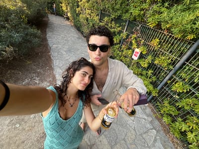 A couple poses for a selfie, leaning in close to each other. The man wears sunglasses and holds two Corona bottles and a purple object. The woman wears a turquoise dress and a necklace. They stand on a stone pathway beside a metal fence, green plants visible through the fence and on the pathway's edge. The photograph is taken at a tilted angle, with the ground and pathway appearing slightly warped.