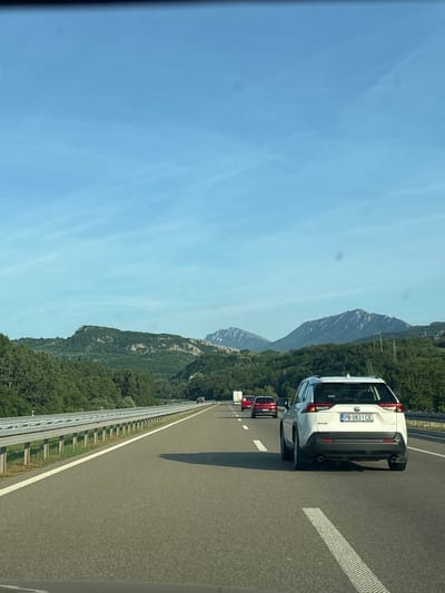 The image shows a highway in Serbia, specifically within the Градска општина Нишка Бања region. A white Toyota RAV4 is prominently featured in the right lane, with the license plate "PB 0831 CE". Other cars, including at least one red vehicle, are visible further ahead in the same lane. The highway is lined with a metal barrier and green vegetation on the side. In the background, mountains are visible under a clear, blue sky.