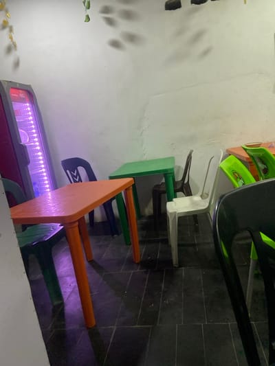 Various colored tables and chairs are arranged in a row. The table colors are orange, green, and green-yellow. The chairs are black, white, green, and blue. A refrigerated display case with pink trim and purple lighting is at the end of the row of tables. The tables and chairs are arranged against a gray tiled floor and white wall with shadows.