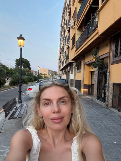 The image is a selfie taken by a blonde woman in Benimàmet-beniferri, Spain. She is standing on a sidewalk next to a street with parked cars. Behind her is a building with balconies and windows. A lamppost is visible to her left. The sky is a light blue, indicating it is daytime.