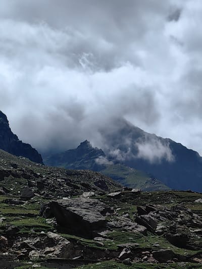 The image depicts a mountainous landscape in Yari-Khoksar, India, under a cloudy sky. The foreground is dominated by a rocky, uneven terrain covered with sparse green vegetation and scattered large boulders. The midground features steep, dark mountain slopes with some patches of green. In the background, larger mountains rise, partially obscured by thick, grey clouds that create a dramatic and moody atmosphere. The overall impression is one of rugged, natural beauty with an imposing, overcast sky. There are no people or discernible man-made structures visible in the scene.