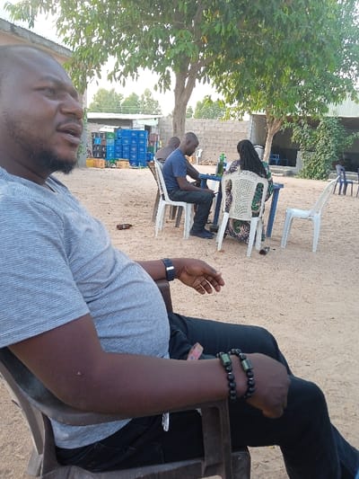 The image captures an outdoor scene in Barama Lokuwa, Nigeria, featuring several individuals in a relaxed setting.
In the foreground, a man is seated in a brown plastic chair, facing left and slightly away from the camera. He has a short beard and is wearing a light grey striped t-shirt, black trousers, a black digital watch on his left wrist, and two beaded bracelets (one black, one green and black) on his right wrist. He appears contemplative, looking into the distance. His posture suggests a moment of rest or observation.
In the mid-ground, under the shade of a large tree with green foliage, two other individuals are seated at a blue plastic table, also on white plastic chairs. One man, wearing a blue t-shirt and dark trousers, is seen from the side, looking down, possibly at a phone. Across from him, a woman with dreadlocks is visible from the back, wearing a colorful patterned dress. A green bottle is on their table, along with another object that might be a phone.
The ground is sandy and dry, indicating an outdoor, possibly unpaved area. In the background, stacks of blue and yellow plastic crates are visible, likely used for drinks or storage, against a concrete block wall. Further back, simple buildings with corrugated roofs can be seen. Another plastic chair is placed randomly to the right. The natural light suggests it is daytime, possibly late afternoon, given the ambient, soft lighting. The overall atmosphere appears casual and social, characteristic of a local outdoor gathering spot or eatery. There is no visible text in the image.