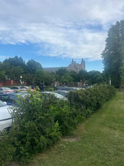 The image shows a parking lot adjacent to a park in Westmount, Canada. In the foreground, there are green bushes and grass. Behind the bushes, several cars are parked. A person is standing near a dark-colored SUV with its trunk open, looking at the contents. In the background, there are trees and a large stone building with multiple towers, which appears to be a church or university. The sky is partly cloudy.