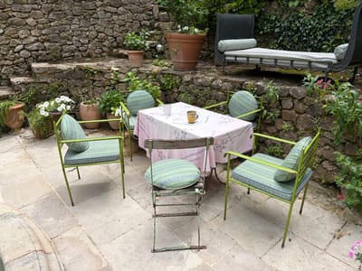 An inviting outdoor patio area in Saint-Victor-la-Coste, France, features stone paving and terraced stone walls covered in lush green foliage. At the center, a round table is draped with a pink and white floral tablecloth, holding a white and a yellow patterned mug, suggesting a recent or upcoming casual drink. Surrounding it are four vibrant lime green metal chairs, each adorned with patterned green and blue striped cushions and round or bolster back pillows. A fifth, older folding chair made of wood and metal, with a matching round green cushion, sits slightly apart from the main grouping.

The stone walls behind the seating area incorporate rustic steps and are dotted with various terra cotta pots containing white flowering plants and other greenery, including ivy and other climbing plants. On a higher terrace to the right, a dark-colored daybed or chaise lounge, possibly wrought iron, is furnished with striped green and blue cushions and bolster pillows, offering another spot for relaxation.

The scene appears tranquil and well-maintained, set during daytime under soft, natural light, suggesting an overcast sky or a gentle time of day. No people are present, but the arrangement implies a space designed for casual gathering, conversation, or enjoying a quiet moment amidst the natural beauty of a traditional European garden. The overall impression is one of rustic charm and comfort. There is no visible text in the image.