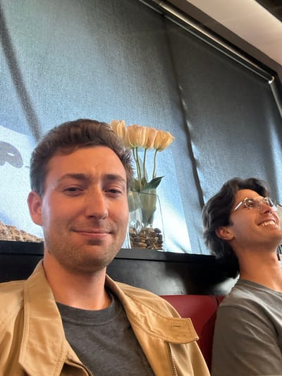 The image captures two males seated in an indoor establishment in Torrance, USA. In the foreground, a male with light skin, short brown hair, and stubble is centrally positioned, looking directly at the camera with a subtle smile. He wears a beige jacket over a gray t-shirt. To his right, partially obscured and slightly behind, a second male with light skin, dark wavy hair, and circular glasses looks upwards and to the right with a broad smile. A red upholstered booth seat is visible in the lower right. Between the two individuals, on a black surface, sits a clear glass vase filled with light-colored artificial tulips and decorative brown pebbles. The background consists of a large window covered by a textured, light blue-gray roller blind, with a faint, indistinct white and light blue graphic visible on the left side of the background. An overhead dark structural element or window frame runs across the top of the image.
