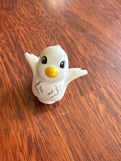 A white plastic duck toy with black eyes, a yellow beak, and grey feather markings on its chest sits on a brown wooden surface. The duck's arms are outstretched. The background is a wooden tabletop with visible grain patterns. The image is taken from a top-down perspective.