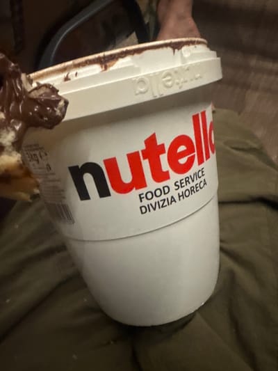 The image shows a large, open container of Nutella, with a portion of it being scooped out with a piece of food, likely a pastry or bread. The container is white with red lettering that says "Nutella" and smaller text below that reads "FOOD SERVICE DIVIZIA HORECA". The lid of the container has some of the chocolate spread on the rim. The container is resting on a green fabric surface, possibly clothing. In the background, a person's feet are visible, and there is a dark object that could be a chair. The context of the image does not provide any information about the location in Miriantsi, Bulgaria.