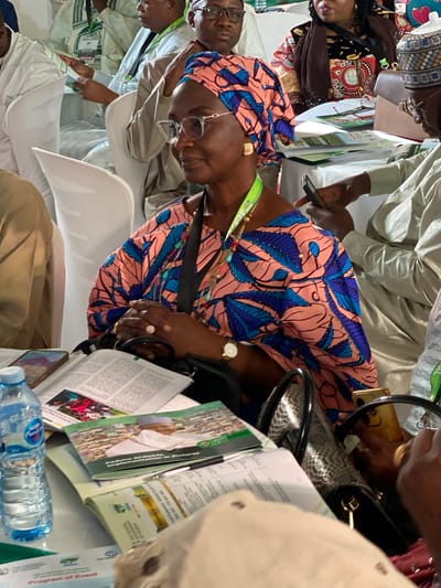 The image shows an indoor scene with several people, likely at an event or conference. One woman is in focus; she wears a colorful patterned top and headwrap, eyeglasses, a wristwatch, and rings. She is looking down at papers. There is a black snakeskin-patterned handbag on the table next to her. There is a water bottle and multiple documents with the word "Kaduna" visible on some of them. Given the presence of "Kaduna" on the documents, it is likely that the location is somewhere in the Nigerian city of Kaduna. The people are dressed in formal attire with lanyards around their necks.