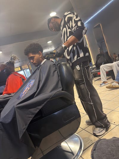This image depicts a barbershop scene in Blouberg, South Africa, where a haircut is in progress. A barber, positioned on the left side of the frame, is actively trimming the hair of a client seated in a black barber chair. The barber wears a striking black and white patterned sweater, dark-wash denim pants with distinctive exposed, frayed seams, and grey and white chunky sneakers. A silver watch adorns his left wrist, and he wears black gloves while operating the hair clippers.
The client, who has dark, curly hair, is draped in a black barber cape featuring a prominent blue and orange geometric design. In the background, another individual with dreadlocks and an orange shirt is partially visible, also appearing to be receiving a service. Further in the background, or reflected in a mirror, a person's legs in light grey sweatpants and patterned slippers are seen.
The setting is a modern, well-lit barbershop with bright overhead strip lighting and several circular ceiling lights. The floor is covered with light-colored tiles. A large mirror reflects parts of the interior, and a silver metal tray rests on the floor near the barber's feet. Faint text, possibly "SALON," is discernible on a wall or mirror in the background. The overall atmosphere is one of focused activity within a professional salon environment.