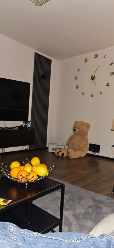 A low-angle shot in a room shows a wall clock, a teddy bear, and a fruit basket. The wall clock has a gold-colored frame and Roman numerals. The teddy bear is brown and appears to be standing on the floor. The fruit basket is filled with oranges and lemons. A television is visible in the background. The location is possibly in Gütersloh, Germany. A person is wearing denim jeans on the right side of the image.