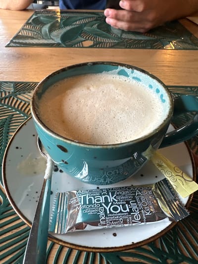 The scene, located in Giebułtów, Poland, depicts a close-up of a table setting. A teal-colored ceramic mug, featuring a speckled pattern and distressed brown rim, holds a frothy, light brown beverage consistent with a latte or cappuccino. The mug rests on a matching speckled white saucer. A spoon with a teal handle and silver bowl is positioned on the saucer. Two sweetener packets are present: a silver foil packet prominently displaying "Thank You" in multiple languages, and a yellow stick-packet with a swirl pattern. These items are situated on a light-toned wooden table, partially covered by a green placemat embossed with a leaf-like pattern. In the upper background, two human hands are visible; one on the right holds a black smartphone.