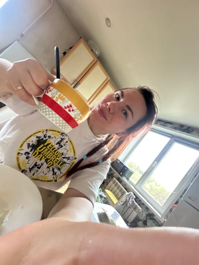 A young woman is taking a selfie in what appears to be a kitchen. She is holding a colorful mug in her left hand, which also has a ring on the ring finger. The woman is wearing a white t-shirt with a "Yellow Umbrella Day" graphic on it. The background includes kitchen cabinets and a window. The precise location of the image within the city of Cheryomushki, Russia, cannot be determined from the details in the photograph.