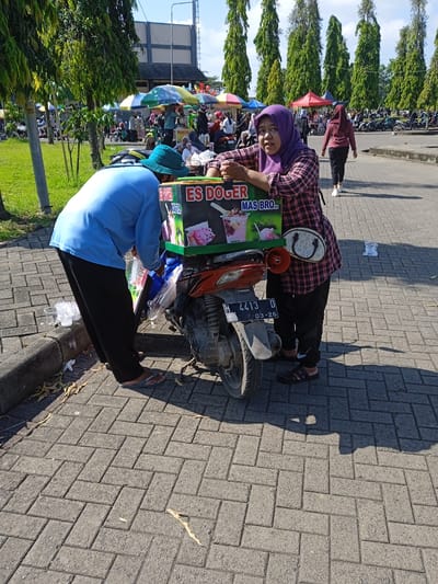 The scene is a street market in Kebondalem, Indonesia. A man in a light blue shirt and a blue bucket hat is preparing "Es Doger" (an Indonesian shaved ice dessert) from a box attached to the back of a parked orange Honda motorcycle with license plate H 4413 D expiring in March 2026. A woman wearing a purple hijab and a pink plaid shirt leans against the "Es Doger" box, seemingly the vendor or assistant. The market stretches into the background with various vendor stalls under colorful umbrellas and numerous parked motorcycles. People are walking around the market, including one woman in a maroon shirt and black pants. Tall green trees line the edge of the parking lot. The ground is paved with gray rectangular bricks. Litter, including plastic cups and bags, is visible on the ground.