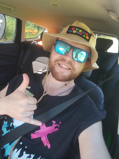 A man is taking a selfie inside a car. He is wearing a tan bucket hat with a colorful geometric patch, blue mirrored sunglasses, and a black t-shirt with a pink abstract design. He has reddish-brown hair and a beard and is smiling. His left hand is giving a thumbs-up. The car interior is visible, including black seats and a seatbelt. The background outside the car window shows green trees. The image is dated July 3, 2023. There is no indication of the location being Zashchita, Russia.