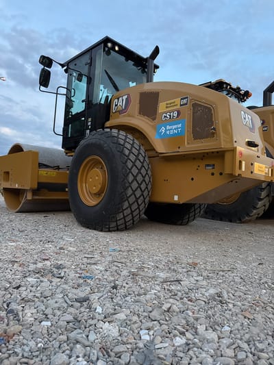 The image shows a close-up, low-angle view of a yellow Caterpillar CS19 soil compactor. The compactor is parked on a surface of crushed stone and debris. The vehicle has a large, ribbed tire on the right side, a smooth drum roller on the left, and a black cab with large windows. The CAT logo is visible on the side of the compactor. A blue and white "Bergerat Rent" sticker is also present. The sky in the background is overcast. The image appears to have been taken in an outdoor industrial or construction setting. The location is identified as Grenay, France.