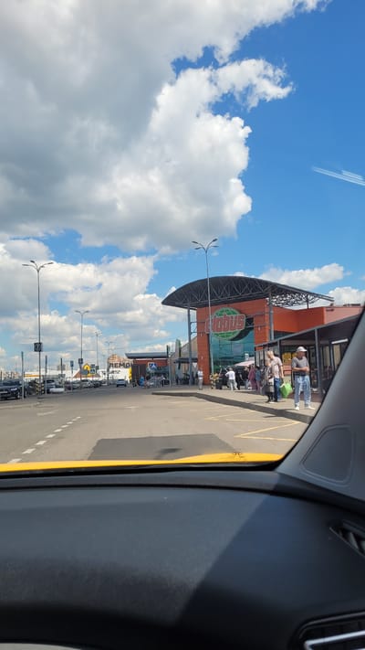 The image, taken from inside a vehicle, shows a street scene in Klimovsk, Russia. The perspective is from a car window, with the dark interior of the car visible in the foreground on the right. The scene outside is dominated by a bright blue sky with fluffy white clouds, suggesting it is daytime. A contrail from an airplane is visible high in the sky.

The main subject is a large orange building with a green and orange sign that reads "Globus" in white lettering. The building appears to be a retail establishment, possibly a supermarket or hypermarket. In front of the building, there is a paved area with yellow lines marking parking spaces. Several people are standing in a line in front of the entrance to the Globus building, some carrying shopping bags. This suggests a queue for entry or service.

A road runs alongside the building, with more parked cars and streetlights visible. The road also has white dashed lines indicating lanes. The overall impression is a sunny, everyday scene at a busy shopping location. The lighting and shadows suggest the sun is high in the sky, likely midday. No specific emotions are discernible from the distant figures.