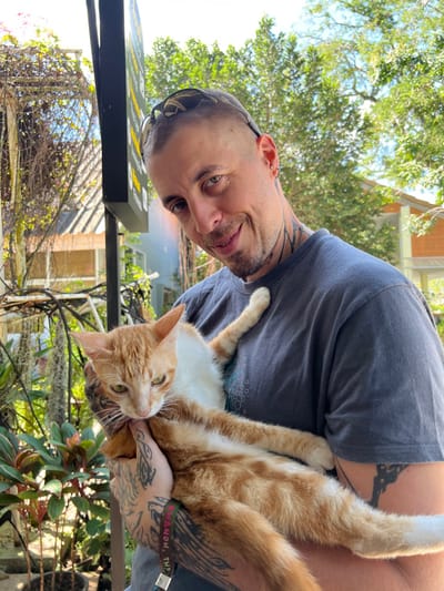 A man holds an orange and white cat. The man has short hair, tattoos on his neck and arms, and sunglasses propped on his head. The cat is held in the man's arms, its legs extended. They are outside, with trees visible in the background. A black sign with yellow lettering is partially visible on the left. The photo is slightly tilted to the side.