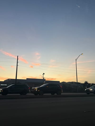 The image captures an outdoor street scene at dusk or sunset in Aventura, Florida. The sky transitions from a light blue at the zenith to an orange-yellow hue at the horizon, marked by scattered, thin, pink-orange clouds.
In the mid-ground, a multi-lane roadway is visible with three vehicles. On the left, a dark-colored SUV is partially visible, appearing to move left-to-right. In the center, a dark Cadillac XT-series SUV, distinguished by its illuminated front lights, moves right-to-left. On the far right, a light-colored SUV is partially visible, also moving right-to-left. All vehicles appear to be in motion.
In the background, silhouetted commercial buildings line the horizon. On the left, a building displays a partial sign reading "HOUSE OF NAILS," confirming the Aventura, Florida location. A tall, dark utility pole with multiple horizontal power lines is positioned on the left side of the frame. On the right, a black street light pole with a single, illuminated yellow lamp stands prominently. Sparse palm trees are visible near the horizon on the far right. No individuals are discernible.