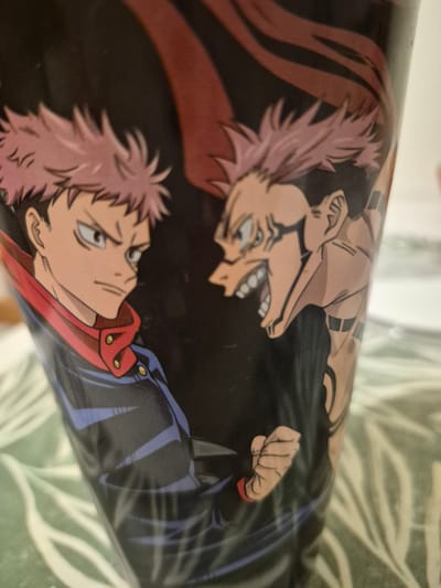 An animated illustration depicts two characters on a black cylindrical object, possibly a cup or container. The character on the left has spiky pink hair, blue eyes, and is wearing a dark blue jacket with a red collar and gold buttons. The character on the right has similar pink hair and appears to be roaring with its mouth open, displaying sharp teeth and a menacing grin. This character also has black markings on its face and arms. A red, ribbon-like element flows from the top of the image behind the characters. The object is placed on a green and white patterned surface, possibly a tablecloth or placemat. The lighting suggests an indoor setting. The exact location is Unknown, Unknown.