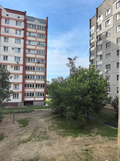 This media file depicts an outdoor scene with two apartment buildings in Zhlobin, Belarus. The building on the left is taller and has a facade with alternating white and reddish-brown sections. It features numerous windows, air conditioning units, and a pointed roofline. The building on the right is a gray, multi-story structure with a simpler, more uniform facade and also many windows.
In the foreground, there is a dirt and grassy area that appears to be a playground. A swing set with two swings is visible, along with part of a slide. A couple of people, one adult and one child, are walking on a paved path between the buildings and the playground.
The scene is set under a bright blue sky with some scattered clouds, suggesting it is daytime. A few tall trees with lush green foliage are present, casting shadows on the ground and obscuring parts of the buildings. Power lines are visible strung between the buildings and across the sky. The overall impression is a typical residential area in a town or city. There is no visible text in the image.