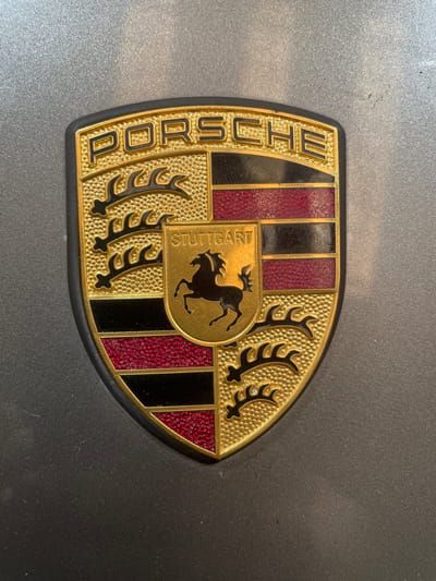 The image displays a close-up, eye-level, frontal view of a Porsche emblem affixed to a gray, textured surface, likely a car. The emblem is shield-shaped and features the Porsche logo with the name "PORSCHE" prominently displayed at the top in gold lettering with black outlines. Below the name, the emblem is divided into quadrants with alternating red and black stripes, separated by gold borders. In the center of the emblem is a smaller gold shield containing a black horse silhouette and the word "STUTTGART" in gold lettering. The surface on which the emblem is mounted appears to be a car's exterior, possibly a hood or trunk, given its metallic gray color and slight texture. There are no people or other objects in the frame. The lighting is even, suggesting an outdoor or well-lit indoor environment. The location in Gerstheim, France is not discernible from the image itself.