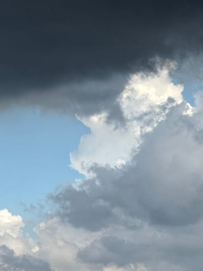 The image captures a dynamic sky with a mixture of dark storm clouds and bright white cumulus clouds. A patch of clear blue sky is visible in the upper left quadrant. The clouds are dense and textured, with varying shades of gray and white, suggesting impending rain or recent rainfall. The scene appears to be an outdoor environment, likely a landscape or cityscape given the context of Đa Lộc, Vietnam. No people or specific objects are identifiable within the frame, focusing solely on the atmospheric conditions. The lighting suggests it could be late afternoon or early evening, with the sun partially obscured by the clouds. The overall impression is one of dramatic weather.