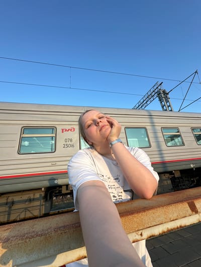 This media file captures a scene at a railway station in Zimovniki, Russia, under a bright, clear blue sky, suggesting either late afternoon or early morning.
A person with blonde hair, wearing a white short-sleeved t-shirt, is positioned towards the right side of the frame. Their left hand is raised, partially obscuring their face, possibly shielding their eyes from the sun or in a contemplative gesture. A silver-colored watch is visible on their left wrist. The person appears to be resting on or leaning against a thick, heavily rusted metal beam or barrier that runs horizontally across the foreground, separating the viewer from the train tracks.
Behind the person and the barrier, a light gray train carriage is prominently featured. The carriage has several windows and a thin red stripe running along its side. Distinct red Cyrillic lettering, "РЖД" (RZD, for Russian Railways), is visible on the carriage, along with the numbers "078" and "23016".
Above the train and against the expanse of the blue sky, elements of railway infrastructure, including overhead power lines and supporting metal structures, are visible. To the far right, a dark paved surface or platform edge can be seen beyond the rusty barrier. The scene depicts a quiet moment at a Russian railway stop.