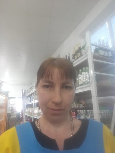 A woman is taking a selfie in what appears to be a store. She is wearing a top with blue and yellow on the shoulder and a black neckline, along with a chain necklace. Behind her are shelves stocked with various items, including what appear to be bottles and glassware. The lighting is bright, and the setting suggests a commercial establishment. The location is Бородулиха ауылдық округі, Kazakhstan.