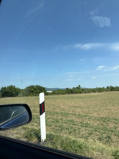 The view from inside a vehicle reveals a rural roadside scene near Pezinok, Slovakia. A car's side mirror is visible in the foreground. A white pole with a black and red band runs vertically beside the mirror. Beyond the pole, a field stretches out, transitioning from green grass to drier vegetation. A tree line marks the far edge of the field. The sky is predominantly blue with scattered clouds.