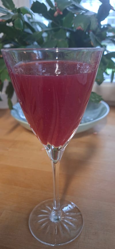 The image shows a martini glass filled with a red liquid, likely a smoothie or cocktail. The glass is positioned on a wooden surface, and a plant with green leaves is visible in the background to the left. To the right of the glass, a light blue bowl or dish can be seen. The lighting suggests it is daytime, and there are no indications of specific weather conditions or location cues beyond Orimattila, Finland, which is provided as context. The overall impression is a still life composition, focusing on the drink and its presentation. There is no visible text in the image.
