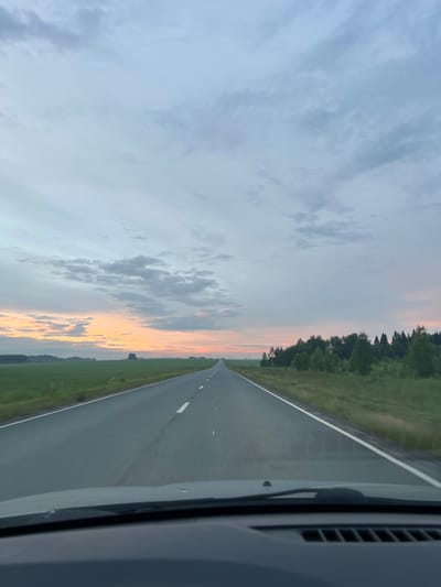 This media file captures a view from a vehicle traveling along a road in Поварёнки, Russia. The scene is dominated by a vast sky, displaying soft pastel colors of a sunset or sunrise, transitioning from pale blue to hues of pink and orange near the horizon. Clouds are scattered across the sky, adding texture to the atmospheric backdrop.
The road itself is paved and appears to be a straight, two-lane highway. White lines mark the edges and center of the road, indicating it is a well-maintained route. To the left of the road, a lush green field stretches out, bordered by a dense line of trees that form a dark silhouette against the colorful sky. To the right, the side of the vehicle is visible, including a windshield wiper and part of the car's interior trim. A road sign, a yellow triangle with a black symbol, is present near the treeline on the left side of the road.
The time of day is clearly either dawn or dusk, given the sky's coloration and soft light. The weather appears calm, with clear skies and no indication of rain or strong winds. The overall mood conveyed is one of peaceful travel and the serene beauty of nature during twilight. The perspective suggests the viewer is a passenger or driver, looking out through the windshield. There is no discernible text within the image.