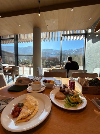 The image shows a breakfast setting with a view of mountains, possibly in or near Озёрное, Russia. Two plates of food are visible: one with an omelet and some chopped fruit, and another with a salad, hash brown, and some dark meat. A cup of coffee and a small bowl of fruit are also on the table, along with a hat. Through the large windows, a mountainous landscape with trees and some structures is visible under a blue sky. Another person can be seen sitting at a table further away by the window.