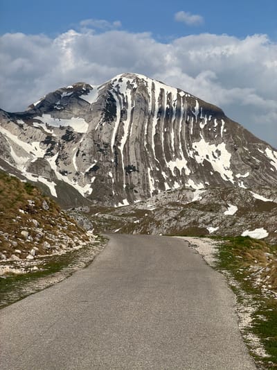 The image depicts a mountainous landscape in Boričje, Montenegro. A large, layered rock mountain occupies the left side of the frame, partially covered with snow patches. The sky above is a mix of blue and cloudy patches. A paved road is visible on the right side of the image, with grassy terrain and rocky patches bordering it. There are no people visible in the scene. The primary focus is on the mountain and the surrounding natural landscape.