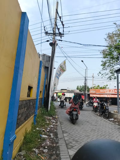 This is a street-level view in Katrungan, Indonesia, on a paved road with a yellow and blue striped wall on the left. The wall is marred with graffiti and is lined with a strip of overgrown grass and debris. Overhead, a complex network of utility wires crisscrosses the sky, supported by a leaning utility pole. Several people on motorcycles are traveling down the road. A large orange sign for "SOTO AYAM LAMONGAN" is visible on the right side of the road, indicating a food establishment. The sky is overcast.