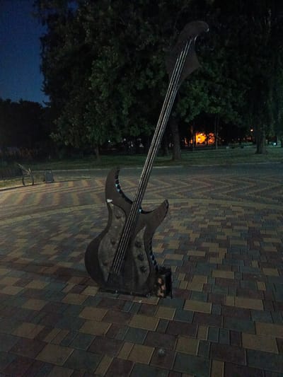 In Biaroza, Belarus, an outdoor scene at night features a large, dark metal sculpture of an electric bass guitar standing upright on a paved walkway. The walkway is constructed of rectangular pavers in varying shades of brown, gray, and green, arranged in a pattern. Behind the sculpture, a dark tree line is visible, with some distant lights emanating from a building with an orange glow. The sky is dark blue, indicating nighttime. The guitar sculpture is detailed, with visible strings and tuning pegs. The base of the sculpture appears to be a small, dark metal box.