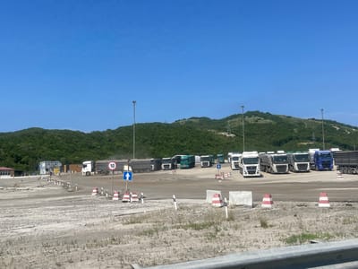 The image shows an outdoor scene in Verkhnebakanskiy, Russia, likely a truck stop or parking area. In the foreground, there is a gravelly, undeveloped area with sparse vegetation and several traffic cones, some with red and white stripes. A metal barrier runs along the bottom of the frame. In the midground, a paved area is occupied by a line of parked semi-trucks. Many of the trucks are white, with some green and a blue one visible. Some trucks are empty cab-over-engine designs, while others are dump trucks. Traffic signs are present, including a circular sign with the number "10" and a blue directional arrow sign pointing upward. A small building with windows and a sign reading "МАГАЗИН" (Shop) is visible to the left, alongside a smaller building with awnings and signage indicating "24/7". In the background, rolling green hills covered in dense trees rise under a clear, bright blue sky. Several tall light poles are scattered throughout the scene. The overall impression is a functional, somewhat industrial area set against a natural backdrop.