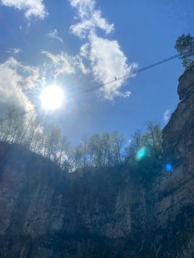 The image depicts a view from below, looking up at a rocky cliff face and a bright, sunlit sky. A suspension bridge or walkway with a linear pattern of platforms extends from the top of the cliff into the sky. Trees are visible along the top edge of the cliff. The environment consists of a rock formation, trees, and the sky with clouds. The location is Kul'metovo, Russia, based on the user's request.
