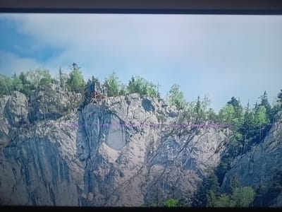 The media file displays a wide, rocky cliff face topped with trees under a bright, partly cloudy sky. The cliff is characterized by its jagged texture and varying shades of gray, indicating a natural, rugged landscape. A small, dark structure, possibly a cabin or observation post, is visible perched precariously on the cliff edge towards the left of the frame. Evergreen and deciduous trees with lush green foliage grow densely on the summit and parts of the slope. The lighting suggests it is daytime, with the sun casting shadows and highlighting the rocky contours. A faint, horizontal purple line appears across the middle of the image, likely a visual artifact. No people, text, or specific events are discernible. The scene conveys a sense of natural grandeur and remote wilderness, hinting at a mountainous or hilly region. Considering the provided location context, this could be a view from a hiking trail or viewpoint in the Oberviechtach area of Germany.